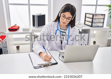Young beautiful hispanic woman doctor having video call writing on document at clinic