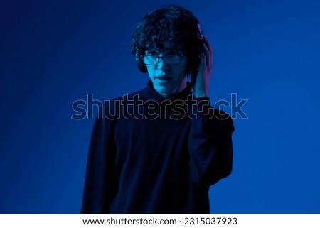 Man teenager wearing headphones listening to music, dancing and singing open mouth smile with glasses, hipster lifestyle, portrait blue background mixed light, fashion style and trends, copying space
