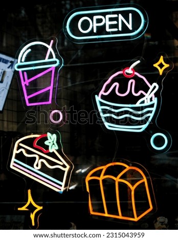 A vibrant neon sign with 'Open' and enticing neon illustrations of cakes, drinks, and breads.

