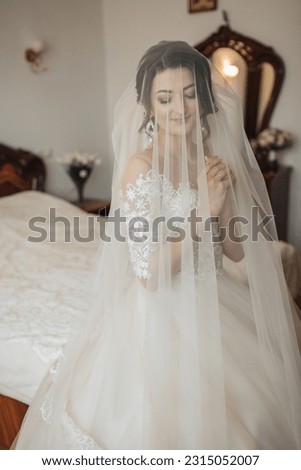 Portrait. A brunette bride in a white dress, wrapped in a veil, looks down and touches her face, poses. Gorgeous make-up and hair. Voluminous veil. Wedding photo. Beautiful bride