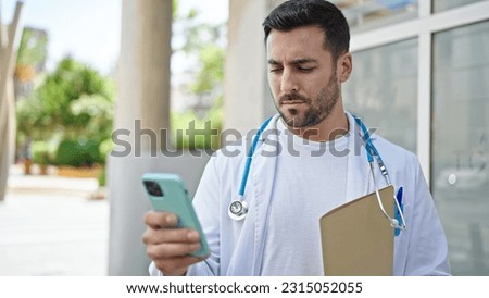 Young hispanic man doctor holding medical report using smartphone at hospital