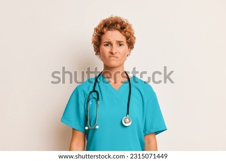 Young female doctor with curly hair and wry face poses at white wall, wearing medical uniform, has stethoscope on her neck, health worker concept, copy space