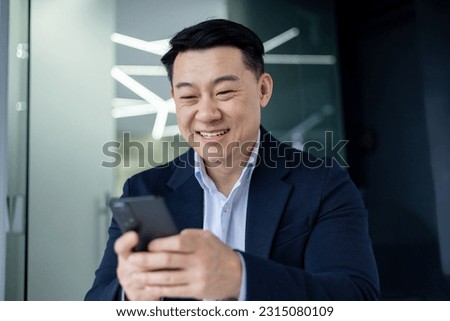 Asian businessman inside the office using the phone, the boss is holding a smartphone in his hands reading news and typing a message smiling..