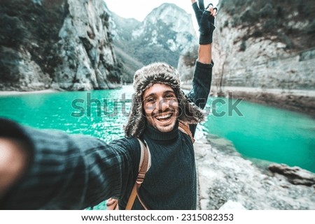 Handsome young man with backpack taking selfie outdoors - Happy hiker standing in front of mountains - Travel blogger enjoying nature view on summer trip 