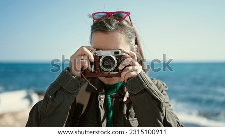 Mature hispanic woman with grey hair tourist wearing backpack taking pictures with vintage camera at seaside