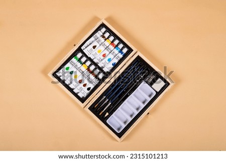 A landscape image of a watercolour paint set in a wooden case on a beige background. The set has a rainbow of colours, paint brushes, a pencil, sharpener and eraser, as well as a pallette.