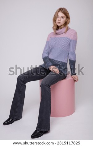 Fashion photo of beautiful elegant young woman in a pretty triple color pink, blue, gray sweater, blouse, black jeans, denim posing on white, soft gray background. Blonde. Model sits on cube, cylinder