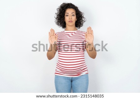 Serious Beautiful pregnant woman standing over white studio background pulls palms towards camera, makes stop gesture, asks to control your emotions and not be nervous