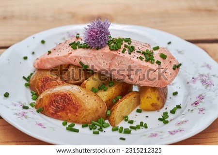 Close up picture on steamed slamon fillet with baked new not pelled potatoes and sprinkled with choped chives and decorated with chives blossom served on rustic style prcelain plate on wooden table.