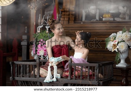 Portrait of beautiful woman in Thai old suit or Thai national dress looks elegant and charming and a little girl sit on ancient elephant seat in old antique Thai houses, Old Thai fashion model concept
