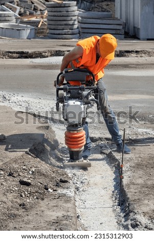 A worker in an orange cape and cap compacts the foundation with a vibratory rammer in a trench on a sunny summer day. Vertical image. Royalty-Free Stock Photo #2315312903