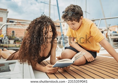 Mother and Son Enjoying a Book on a Boat Deck - A beautiful woman with curly hair, Mediterranean, shares a special reading moment with her pre-adolescent son on the deck of a peaceful boat. Royalty-Free Stock Photo #2315359041