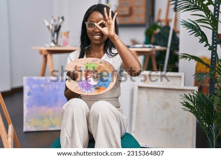 African young woman holding painter palette smiling happy doing ok sign with hand on eye looking through fingers 