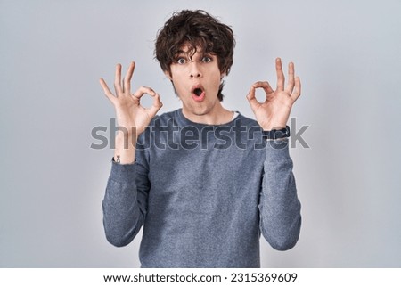 Young man standing over isolated background looking surprised and shocked doing ok approval symbol with fingers. crazy expression 