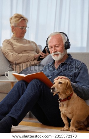 Senior couple spending leisure time at home with their dog. Elderly spouses reading book, listening to music, podcast or watching a movie while sitting on sofa. Happy retirement concept.