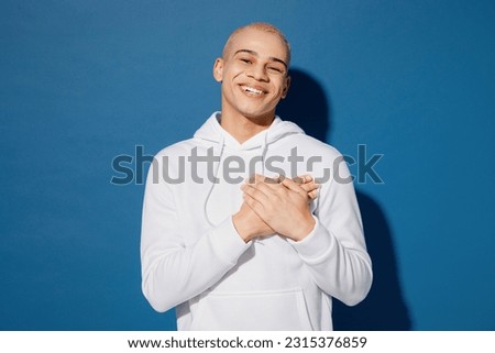 Young thankful smiling happy fun cool cheerful dyed blond man of African American ethnicity wear white hoody put folded hands on heart isolated on plain dark royal navy blue background studio portrait
