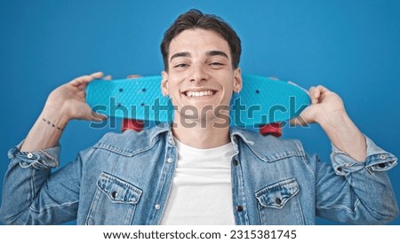 Young hispanic man smiling confident holding skateboard over isolated blue background
