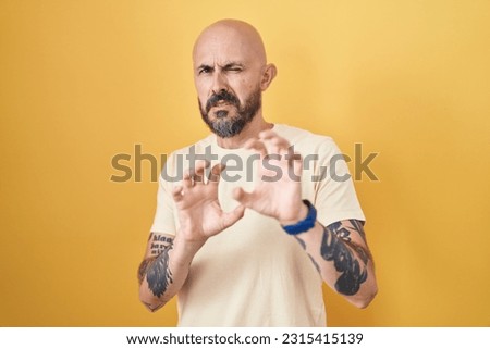 Hispanic man with tattoos standing over yellow background disgusted expression, displeased and fearful doing disgust face because aversion reaction. 