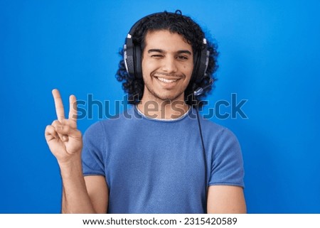 Hispanic man with curly hair listening to music using headphones smiling with happy face winking at the camera doing victory sign. number two. 