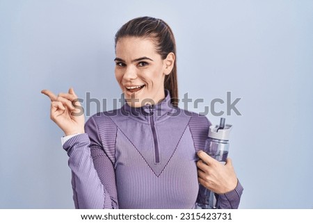 Beautiful woman wearing sportswear holding water bottle with a big smile on face, pointing with hand finger to the side looking at the camera. 