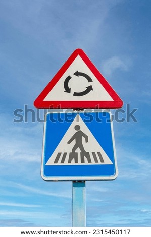 Roundabout and pedestrian crossing road signs against a blue sky background.