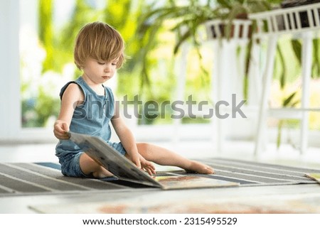 Cute toddler boy reading a childrens book on the floor at home. Daytime care creative activity. Kids having fun with toys. Educational learning games. Family leisure indoor.