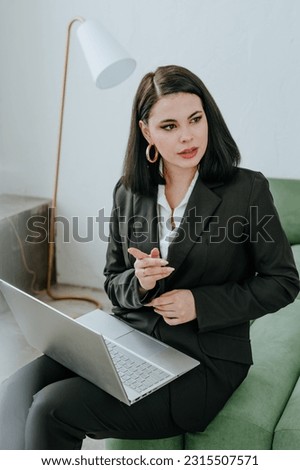 Smiling young businesswoman in a suit with a laptop on her lap siting on a green sofa talking. In the background is a white wall and a white floor lamp.