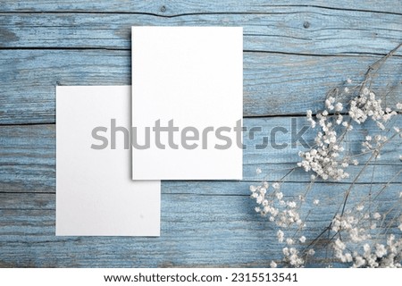 Card mockup, white blank wedding invitation with floral decor on bue wooden background. Greeting card mockup with white dried flowers on table