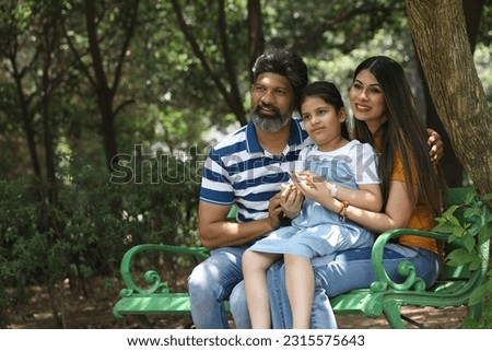 happy Indian healthy family sitting in a city park bench having a cheerful time together. They are surrounded with greenery and serene atmosphere in peaceful and calm environment. Royalty-Free Stock Photo #2315575643