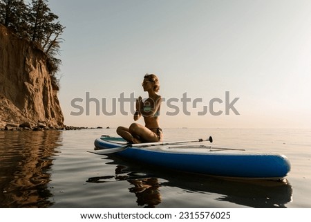 Young woman sitting in lotus pose practising yoga on sup board in quiet sea. Morning or evening meditation.