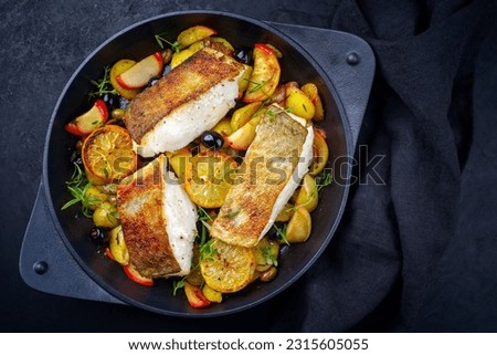 Modern style traditional fried skrei cod fish filet with fried potato, fruits and olives served as top view in a design cast iron pan 