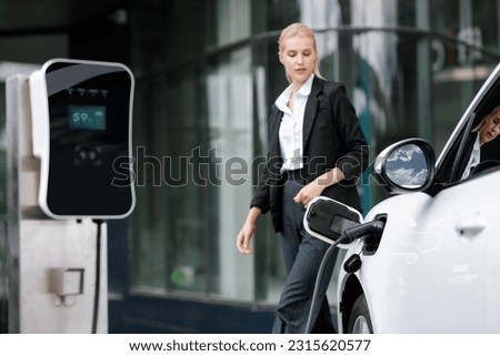 Focus public EV charging station for electric car with blurred progressive businesswoman walking in background. Electric car driven by clean and sustainable energy for ecological concern.