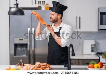 Handsome senior man in cook apron and chef hat cooking fish salmon seafood in kitchen. Portrait of middle aged man cooking seafood in kitchen. Millennial man preparing raw fish salmon and seafood.