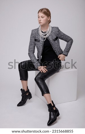 Fashion photo of a beautiful elegant young woman in a pretty suit, black leather pants, trousers, gray jacket, blazer, top posing over white, soft gray background. Model sits on cube. Blonde