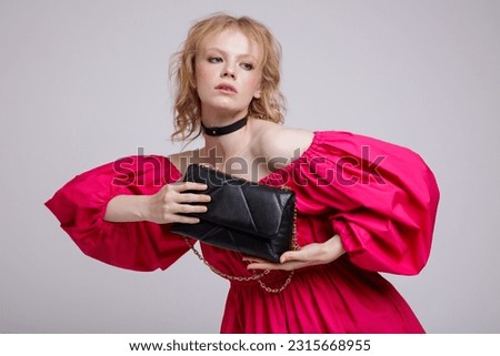 High fashion photo of beautiful elegant young woman in a pretty fuchsia pink dress, black handbag, clutch, choker posing over white, soft gray background. Studio Shot, portrait. Blonde