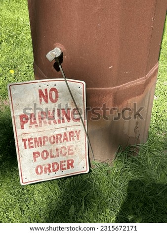 No Parking.  Temporary Police Order.  Dirty, well worn sign at base of post surrounded by grass.