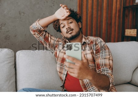 Young Indian man wears casual clothes put hand on head hold use mobile cell phone sits on grey sofa couch stay at home hotel flat rest relax spend free spare time in living room indoor. Lounge concept