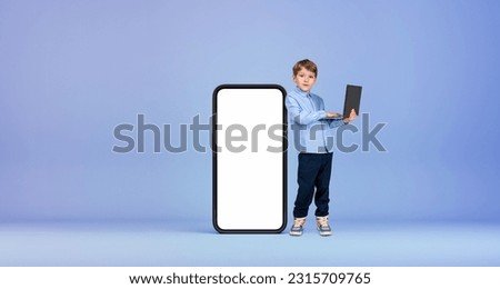 Serious little boy kid with laptop standing near big smartphone with mock up screen over blue background. Concept of advertising, social media and e-learning