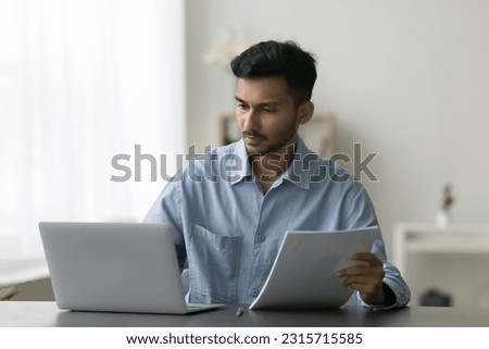 Serious young Indian entrepreneur man reviewing paper documents at work table, working at laptop, looking at screen, checking contract, agreement, consulting online legal service
