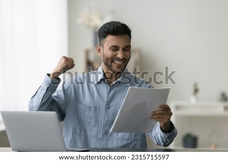 Positive Indian professional man celebrating business success, achievement, victory, holding paper document, letter, notice, contract, agreement, offer, making winner yes hand Royalty-Free Stock Photo #2315715597