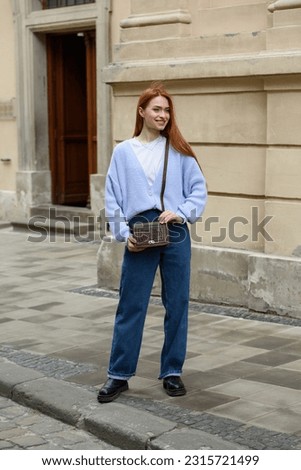 a red-haired girl in a blue jeans and a sweater poses outside with a small leather handbag Royalty-Free Stock Photo #2315721499