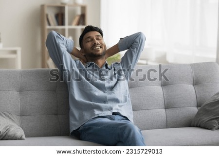 Positive relaxed young Indian man sitting on home sofa, stretching body, leaning on soft back, touching neck with closed eyes, enjoying comfort, coziness, breathing fresh air Royalty-Free Stock Photo #2315729013