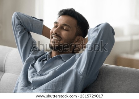 Happy relaxed handsome Indian guy sitting on comfortable sofa, leaning on back with head on hands, smiling with closed eyes, breathing fresh cool air, enjoying calm leisure at cozy own home Royalty-Free Stock Photo #2315729017