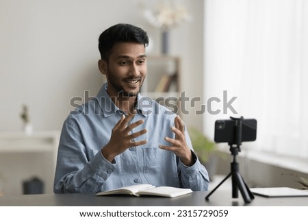 Cheerful excited young Indian blogger recording selfie video on mobile phone, speaking at cellphone fixed on tripod, smiling, laughing, using hand gesture, enjoying self shooting Royalty-Free Stock Photo #2315729059