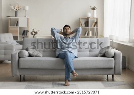 Serious calm young Indian man resting on home couch, sleeping, enjoying leisure time, silent peaceful break, pause, relaxing, sitting on comfortable sofa in modern cozy apartment