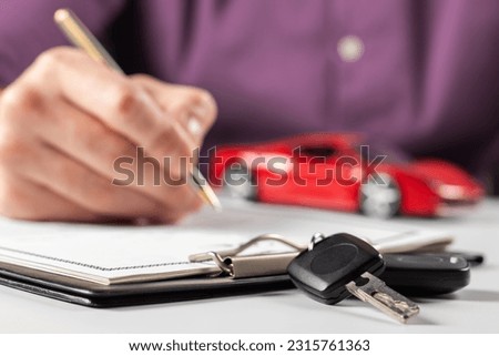 Man signing car insurance document or lease paper. Writing signature on contract or agreement. Buying or selling new or used vehicle. Car keys on table on red car background. Warranty or guarantee.