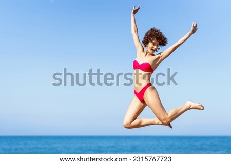 Smiling carefree young female with curly red hair in pink bikini, raising arms and jumping with excitement in air while looking down in daylight against blurred sea and blue sky