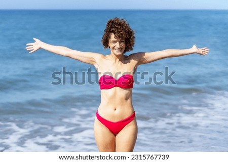 Cheerful young curly ginger haired female in pink bikini with outstretched hands and looking at camera, while standing near foamy rippling seawater against waving sea and blue sky