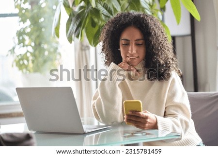 Young happy smiling pretty latin woman holding smartphone using cellphone modern technology, looking at mobile phone while remote working or hybrid learning on laptop at home sitting at table.