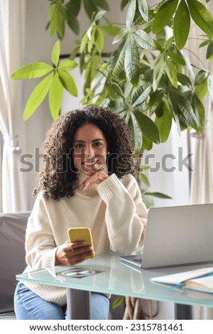Young happy smiling pretty curly latin woman holding smartphone using cell device modern technology, looking at camera with mobile phone in hand sitting at table a home, vertical shot.